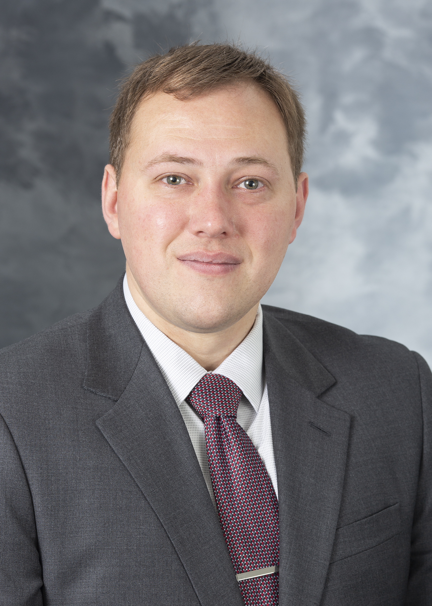 Travis Grimm, smiling light complexion man with brown hair, wearing a grey suit and patterned tie over a white dress shirt.