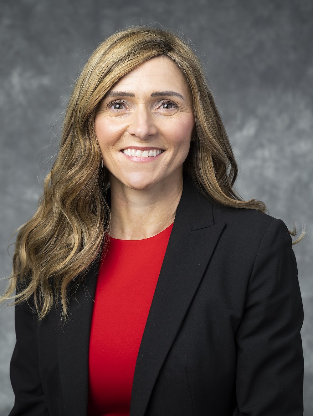 Nancy Lynch, smiling Caucasian woman with long, wavy, dark blonde hair wearing a red shirt and black blazer.