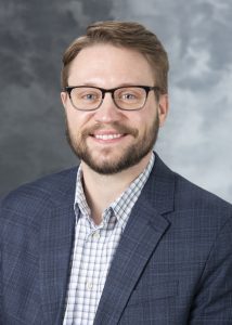 Jack Talaska, smiling man with white skin, short, parted hair, a close cropped beard, and dark framed glasses, wearing a blue and white collared shirt under a blue sport coat.