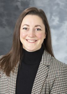 Taylor Buresch, Smiling woman with brown hair, wearing a black mockneck and houndstooth check patterned blazer.