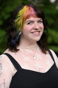 Emily Myers, smiling woman with colorfully highlighted hair wearing a black shirt with straps over a lace layering top.