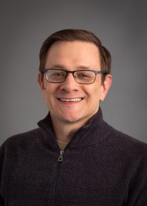 Mark Whittaker, a smiling man with brown hair and glasses wearing a dark grey sweater.