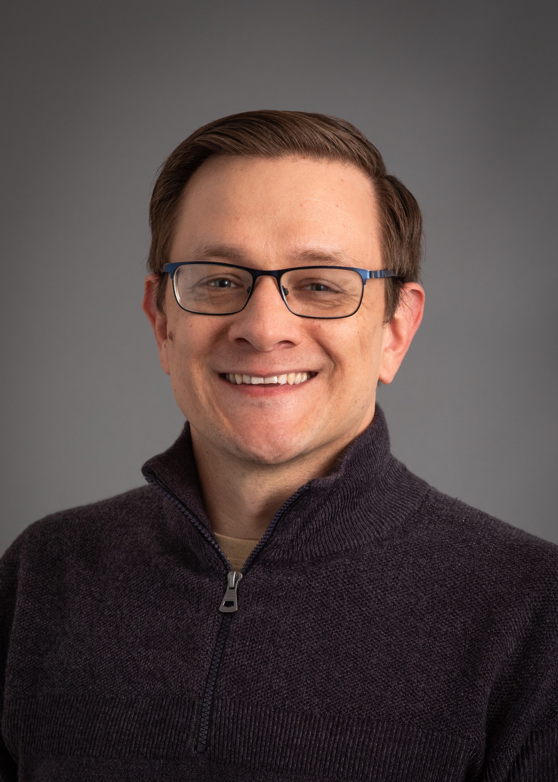 Mark Whittaker, a smiling man with brown hair and glasses wearing a dark grey sweater.