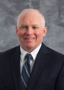 Matthew Valley, smiling man with white hair in a dark blue suit with a white dress shirt and a blue and grey striped tie.