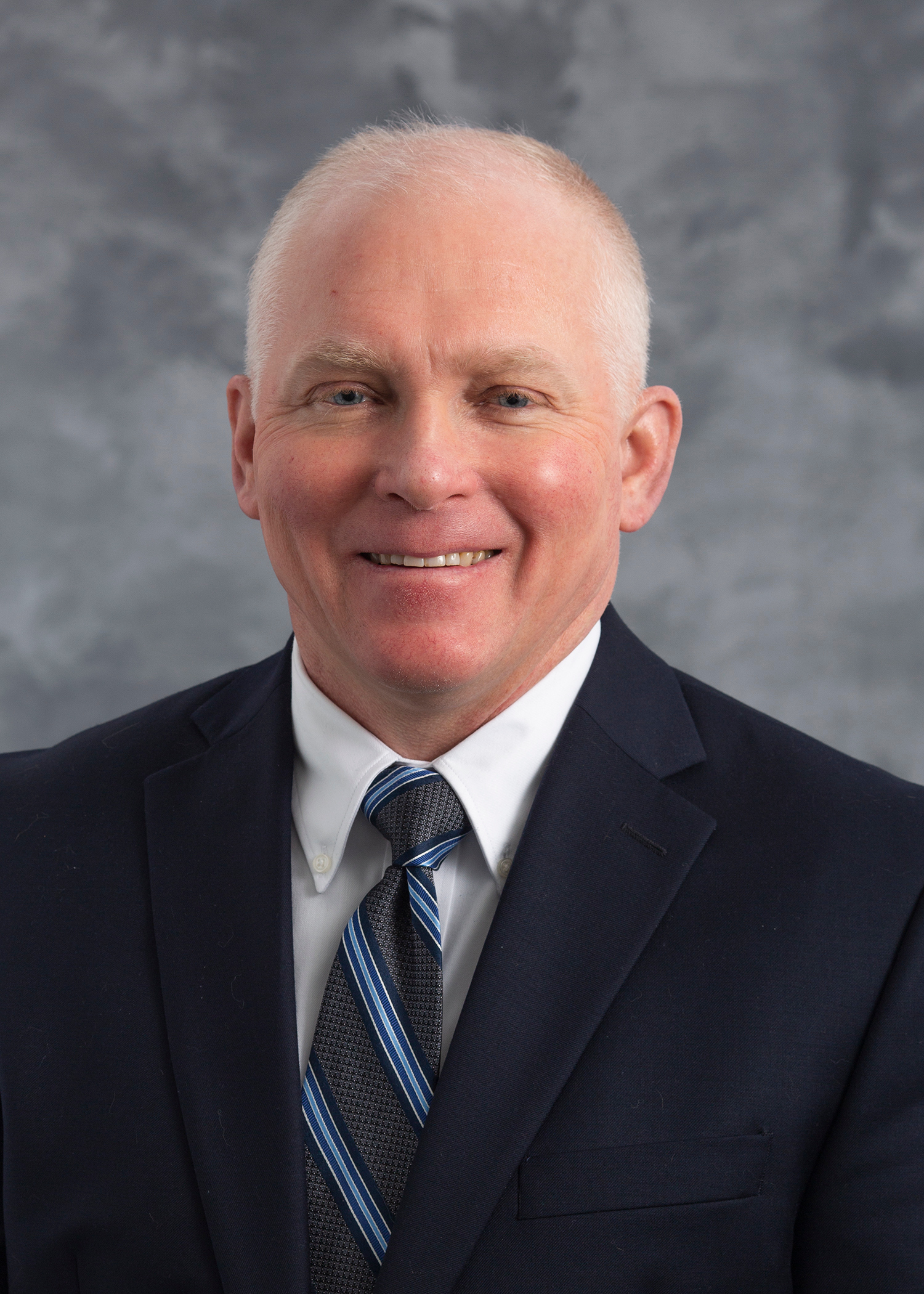 Matthew Valley, smiling man with white hair in a dark blue suit with a white dress shirt and a blue and grey striped tie.