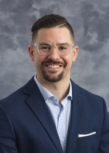 Adam Blaser, smiling man in clear-framed glasses wearing a navy blue blazer over a light blue dress shirt.