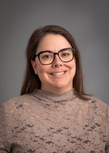 Jen Van Roy, smiling woman with brown hair and black glasses wearing a light brown mockneck sweater.