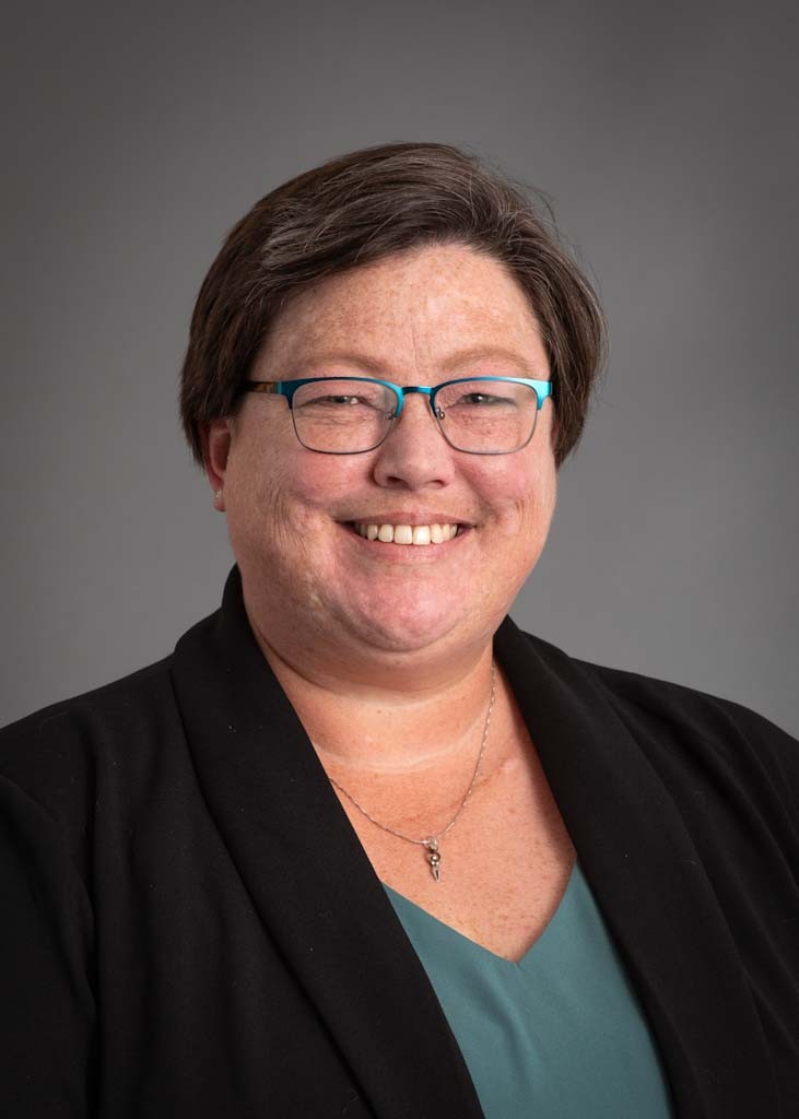 Jennifer Tibbetts, smiling woman with glasses wearing a black blazer over a green blouse.
