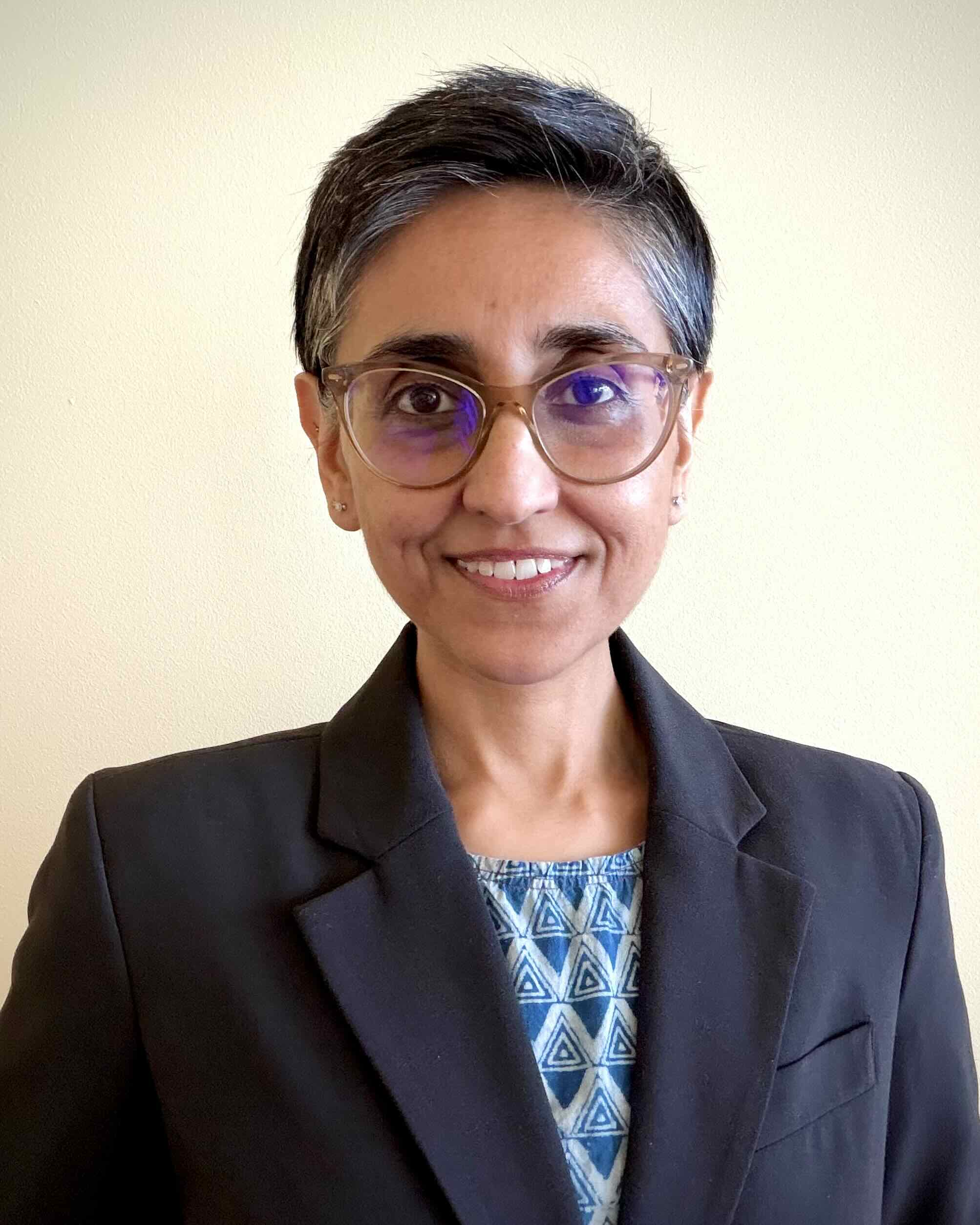 Woman with short dark hair smiling, wearing glasses and a blue shirt with a black jacket.