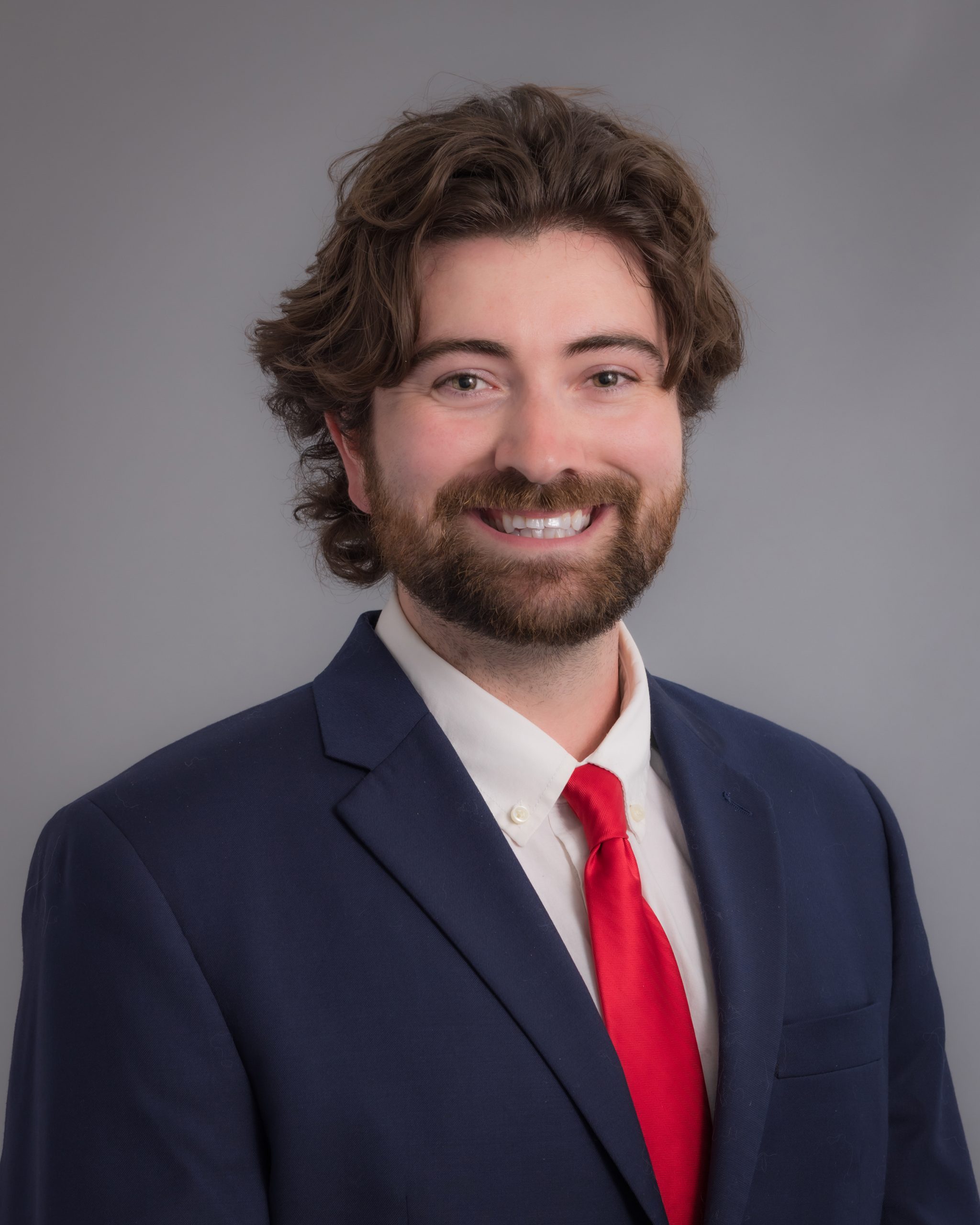 Alex McKenna, smiling man with brown hair and a beard wearing a blue suit over a white dress shirt and red necktie.