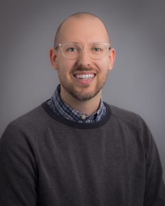 Mitchell Sabez, smiling man with light brown hair and glasses wearing a grey sweater over a blue dress shirt.