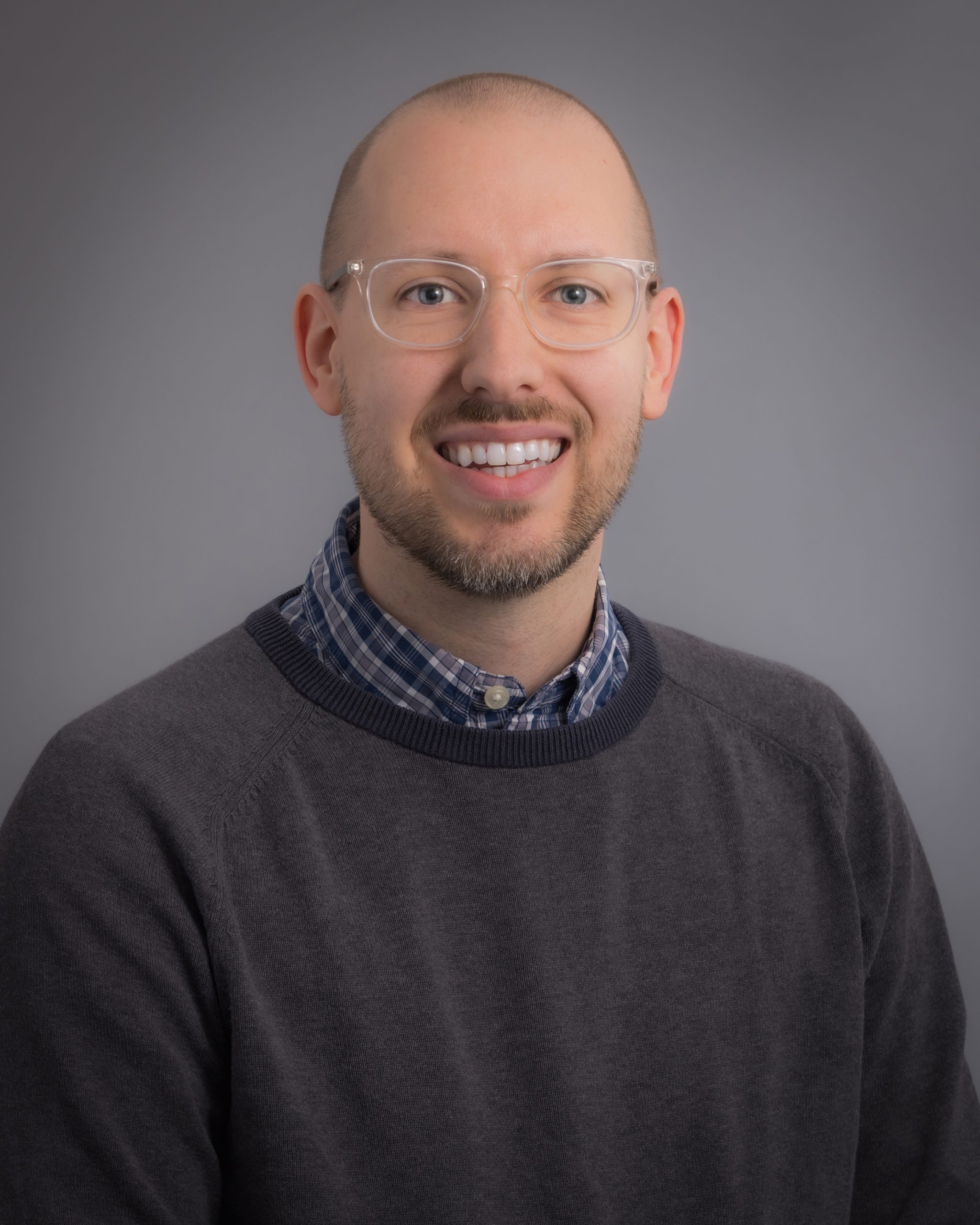 Mitchell Sabez, smiling man with light brown hair and glasses wearing a grey sweater over a blue dress shirt.